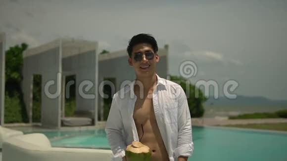 在阳光明媚的日子里穿着无扣白色衬衫和太阳镜在室外游泳池散步的亚洲年轻人异国情调的酒店视频的预览图