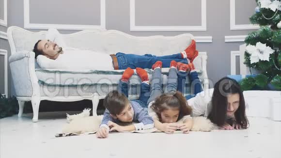 两个孩子和妈妈把腿放在沙发上胳膊肘放在地板上看着摄像机爸爸躺在沙发上视频的预览图