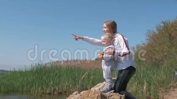 年轻快乐的女性和小男孩望着远方手伸向远处的湖站在水面上的石头上视频的预览图
