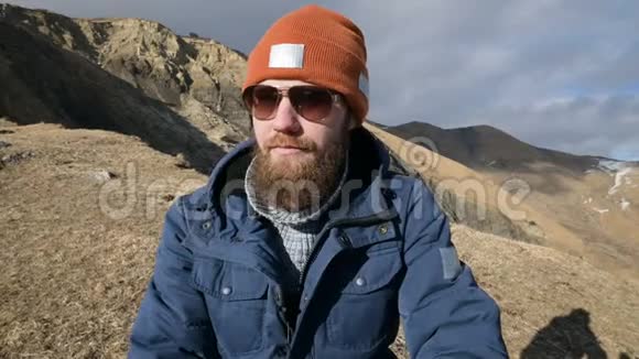 一张戴着太阳镜和帽子的留胡子旅行者的肖像坐在山峦背景下的岩石上大笑视频的预览图
