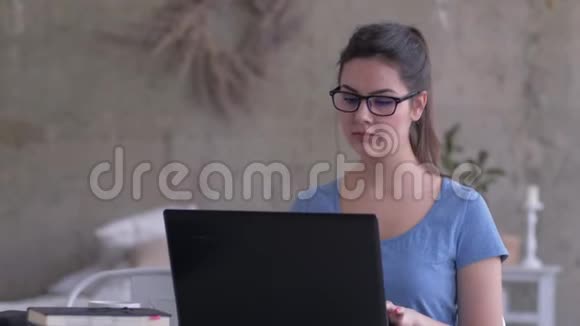 电子学习戴眼镜的现代女孩看笔记本电脑屏幕并在家里的特写键盘上输入文字视频的预览图