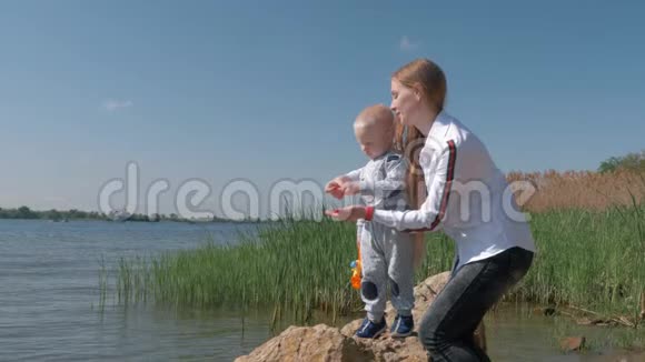 户外游戏年轻的母亲和小儿子在假期里站在河岸上把小石子扔进水里视频的预览图