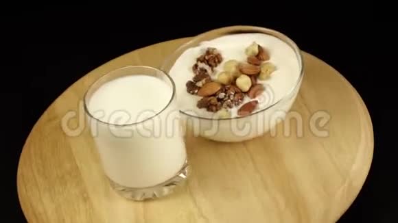 带坚果的酸奶和一杯牛奶在黑色背景的木制平台上旋转视频的预览图