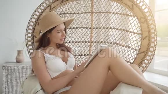 网上购物戴草帽的性感女人坐在舒适的扶手椅上一边拿着银行卡一边通过平板电脑上网购物视频的预览图