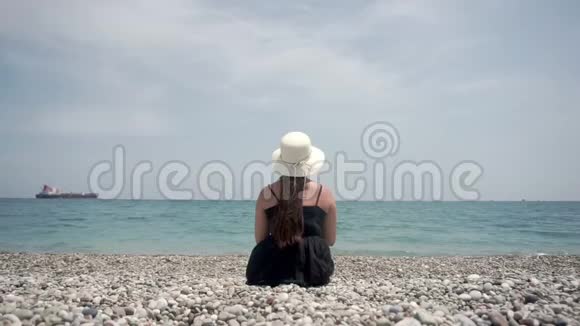 旅游女孩坐在海滩上看着远处某个地方的一艘大货船视频的预览图