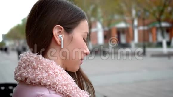 可爱的少女正在通过无线白耳机听音乐秋天在街上的长椅上视频的预览图