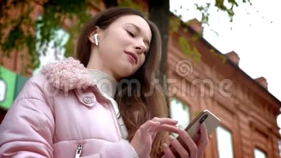可爱的少女正在通过无线白耳机听音乐秋天在街上的长椅上视频的预览图