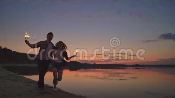 年轻快乐的多民族夫妇手持燃烧着闪闪发光的蜡烛日落时在海边奔跑慢镜头视频的预览图