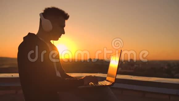 一个穿着帽衫的男人坐在屋顶上听音乐耳机在笔记本电脑键盘上打字视频的预览图