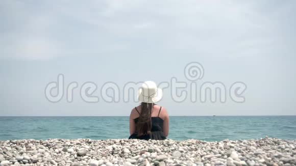 一个戴着宽边帽子的旅游女孩坐在温暖的海洋的岸边欣赏美丽的水景视频的预览图
