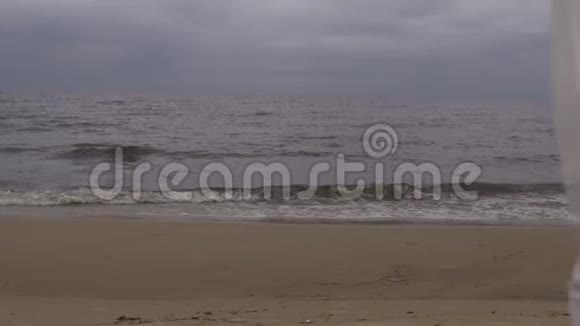 后景美丽的年轻金发女子穿着白色连衣裙朝海边的水上海滩走去在海边的海浪中视频的预览图