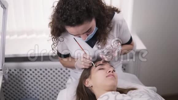 专业美容柜中的美容师正在用棕色染料在年轻女子的眉毛上涂上颜料上色视频的预览图