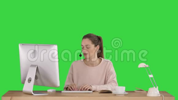 漂亮的年轻休闲女士戴着耳机在绿色屏幕上的电脑上工作Chroma键视频的预览图