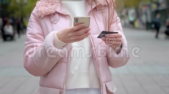 一个年轻的女孩站在一条城市街道上持有智能手机和银行卡通过信用卡进行电子支付视频的预览图