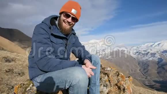 一张戴着太阳镜和帽子的留胡子旅行者的肖像坐在山峦背景下的岩石上大笑和视频的预览图