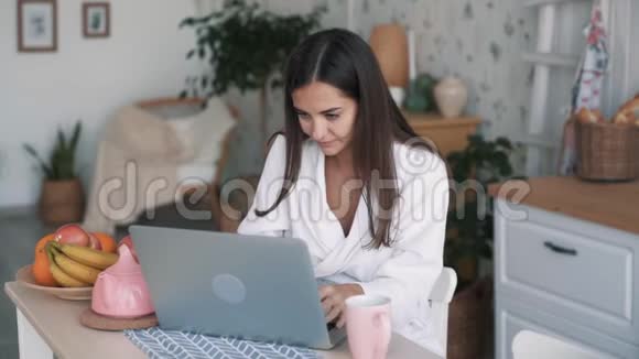 穿着浴袍的女孩在家里用笔记本电脑喝咖啡或茶慢动作视频的预览图