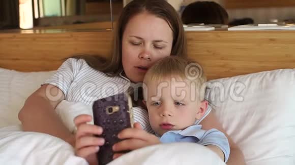 妈妈和儿子躺在毯子下面的床上在电话里玩游戏视频的预览图