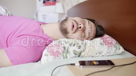年轻的男性穿着粉红色的t恤用枕头睡在床上当手机铃声响起时醒来视频的预览图
