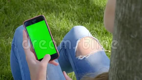 年轻漂亮的女孩穿着公园里的街服撕破牛仔裤他手里拿着一部带有绿色屏幕的智能手机视频的预览图
