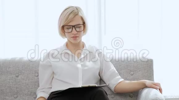 一位年轻漂亮的金发女人戴着眼镜坐在一间浅色公寓的沙发上工作累了脖子疼视频的预览图