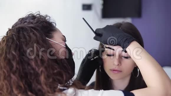 白种人美容师用棕色染料在年轻女子的眉毛上涂上深色颜料从肩膀上拍摄下来视频的预览图