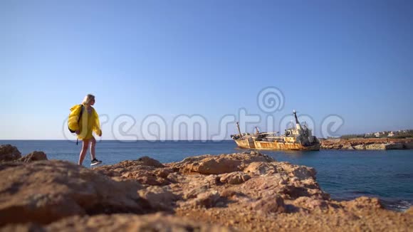 美丽的年轻女游客穿着黄色雨衣在风景如画的大海背景下走在岩石海滩上老了视频的预览图