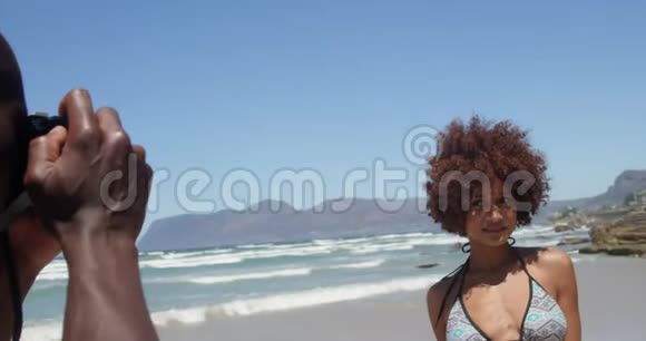 美国黑人青年男子在海滩上用数码相机拍摄女子照片的侧景4k视频的预览图