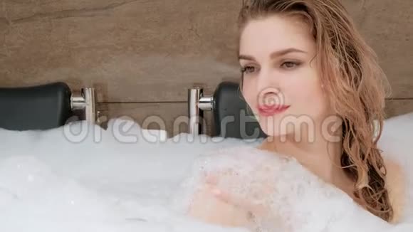 美丽的金发年轻女子躺在浴缸里泡沫休息和放松视频的预览图