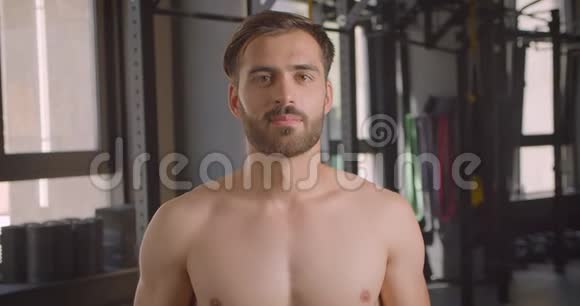 赤裸上身肌肉发达的白种人的特写镜头他正积极地看着摄像机站在室内的健身房里视频的预览图