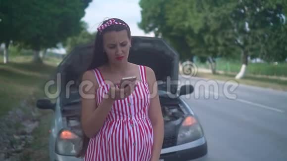 一名怀孕的年轻妇女站在一条乡村道路上的一辆失事汽车附近心烦意乱的女孩打电话寻求帮助视频的预览图
