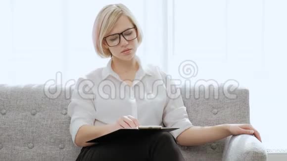 一位年轻漂亮的金发女人戴着眼镜坐在一间浅色公寓的沙发上工作累了脖子疼视频的预览图