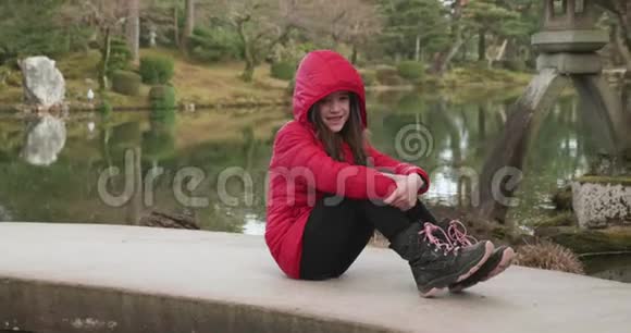 日本金泽的一个日本花园里一个穿着红色夹克坐在桥上微笑拍照的年轻女孩视频的预览图
