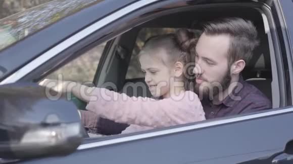 小女孩坐在爸爸身上她的膝盖在车里紧紧地抱着轮子孩子正在学习开车微笑视频的预览图