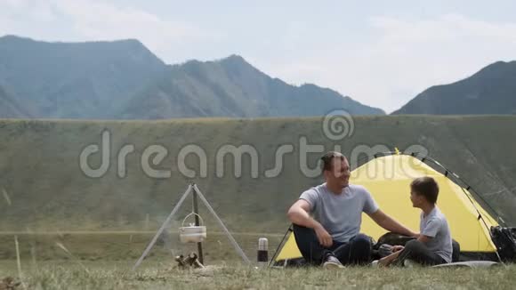 快乐的父子俩坐在帐篷附近篝火旁笑着野营视频的预览图