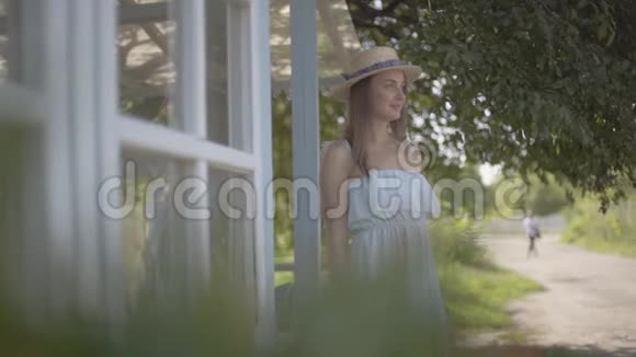 可爱的年轻女子戴着草帽穿着白色连衣裙微笑着在浇灌罐前嗅着野花视频的预览图