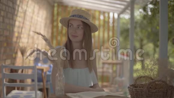 漂亮的年轻女孩戴着草帽和白色连衣裙坐在户外的小木桌上看书农村视频的预览图
