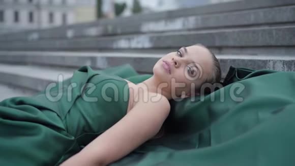 肖像可爱的年轻女孩穿着一件漂亮的晚绿色连衣裙躺在摩天大楼附近空荡荡的城市广场的楼梯上实际视频的预览图
