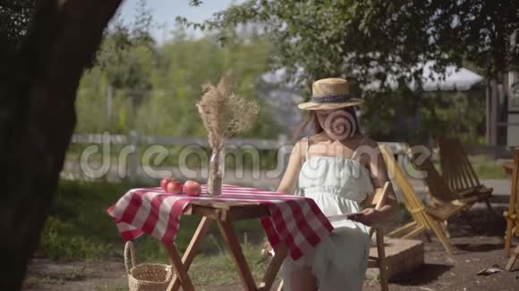 年轻的农村女孩戴着草帽穿着白色连衣裙坐在小桌旁桌上放着花瓶上面放着装饰性的草上面写着视频的预览图