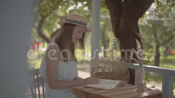 一个年轻漂亮的农村女孩戴着草帽穿着白色连衣裙坐在户外的小木桌旁看书视频的预览图