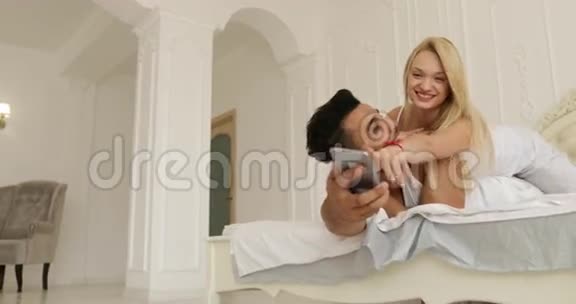 情侣躺床使用手机混搭男人女人开心玩拥抱微笑早间卧室视频的预览图
