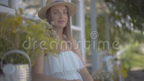 迷人的年轻女子戴着草帽和白色的连衣裙坐在小村房子前面农村生活方式慢慢视频的预览图