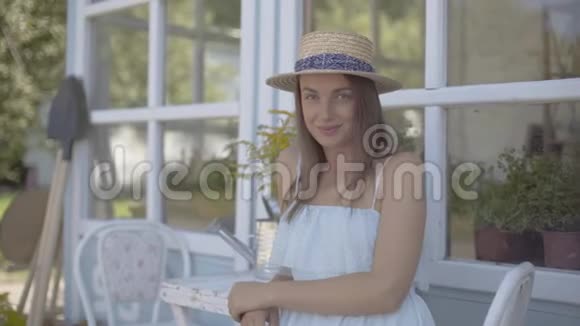 穿着草帽和白色连衣裙的时尚年轻女子微笑着坐在浇花的罐子里嗅着野花视频的预览图