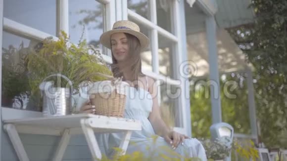 戴着草帽和白色裙子的迷人年轻女子微笑着坐在浇花的罐子里嗅着野花视频的预览图