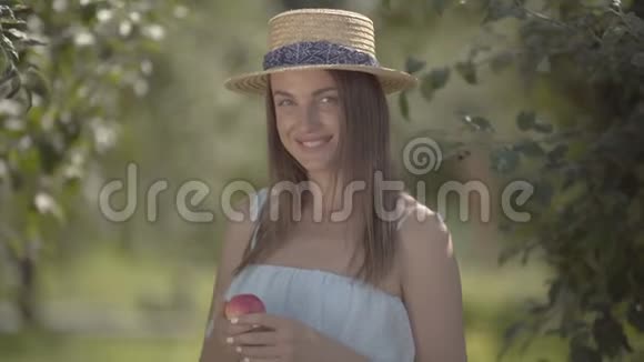 迷人的年轻积极的女人戴着草帽和白色连衣裙看着相机微笑着抱着苹果站在里面视频的预览图