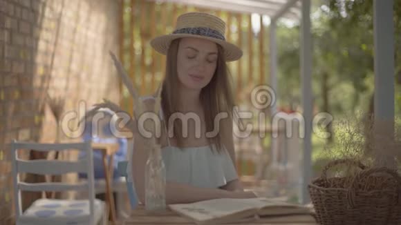 一个年轻漂亮的农村女孩戴着草帽穿着白色连衣裙坐在户外的小木桌旁看书视频的预览图