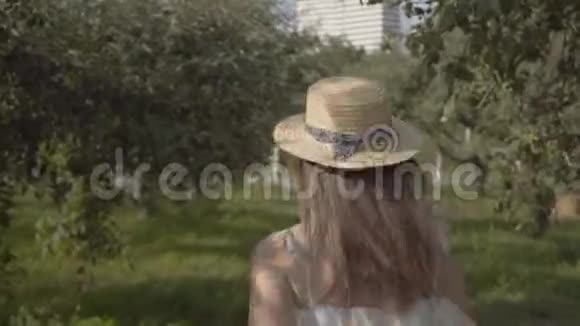 在绿色的夏日花园里一位戴着草帽和白色长裙的迷人年轻女子的后景免费视频的预览图