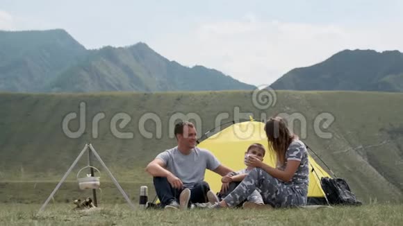 一个幸福的家庭和一个孩子在帐篷前休息喝着热水瓶里的茶概念徒步旅行视频的预览图