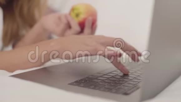 一个年轻的白人女性在笔记本电脑上打字的特写镜头另一只手拿着红苹果的女人视频的预览图