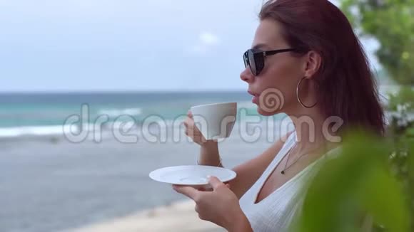可爱的黑发女郎戴着太阳镜和白色连衣裙在海洋附近喝着晨咖啡欣赏着美妙的景色视频的预览图