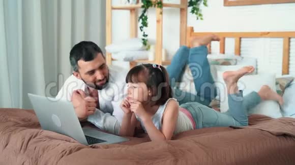 爸爸和可爱的小女孩在家看笔记本电脑上的卡通片躺在床上慢动作视频的预览图